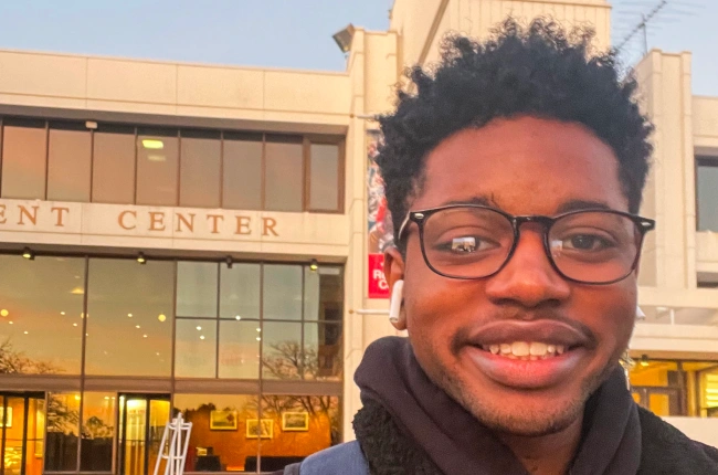 A young Black man takes a selfie in front of a large modern building.