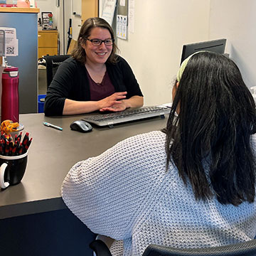 A smiling staff member with dark hair and glasses counsels a female student sitting in front of her in the Office for Student Financial Services.