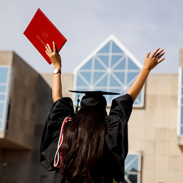 A student facing University Library, arms raised in V, holds her diploma in the air. She is wearing a graduation cap and gown.