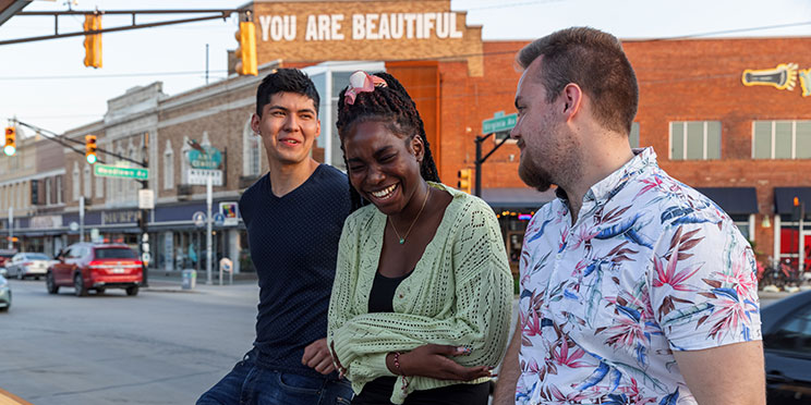 Three students lean against a rail in the Fountain Square neighborhood of Indianapolis. The woman in the middle is laughing. In the background, the top of a orange brick building reads "You Are Beautiful"