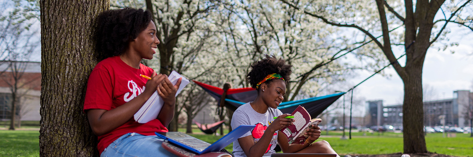 Two female students study on the IU Indianapolis outside between some trees. Someone has hung a hammock behind them. The student on the left is holding a  notebook and has a laptop on her lap. The woman on the left has a book in her hands.