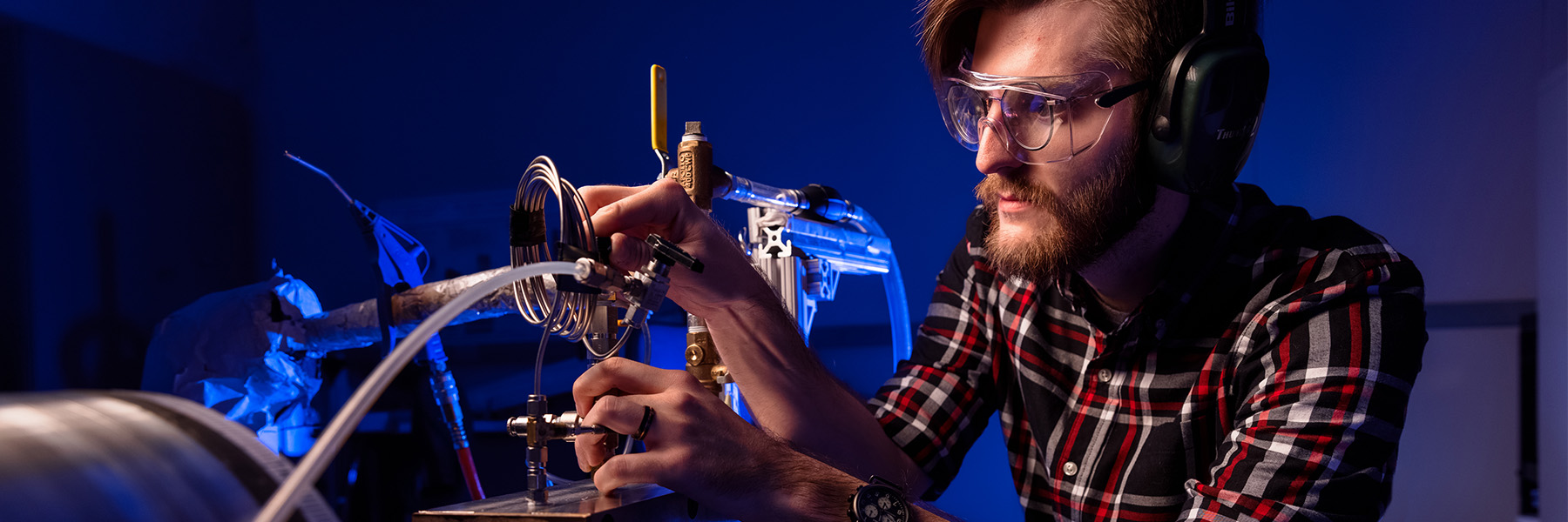 A focused man wearing safety glasses and a headset is diligently working on equipment in a laboratory setting. 