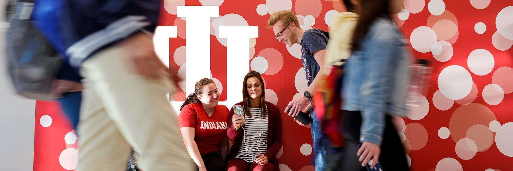 Three students are hanging out in front of a crimson wall with lots of bubbles as other students pass by quickly. The female student in the middle is showing the other two something on her phone. 