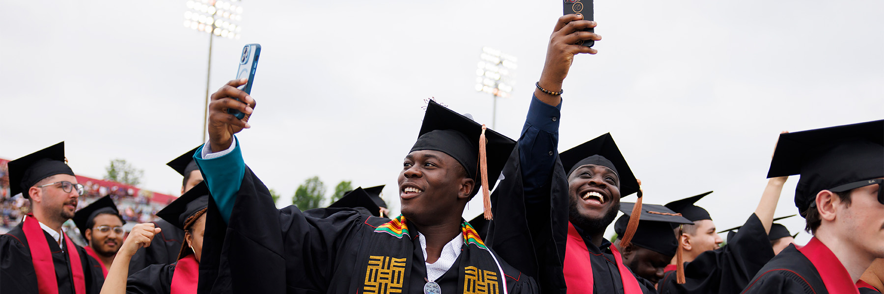 Two excited graduating students take a moment during commencement to hold their phones up and get a picture of the crowds.