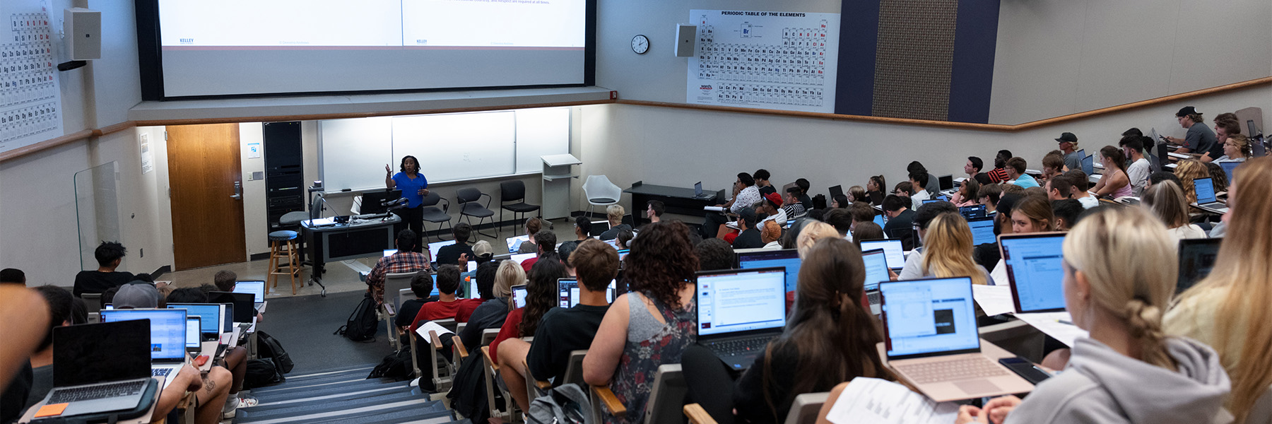 In the lecture hall, students are attentively taking notes on their laptops as the professor teaches the course.