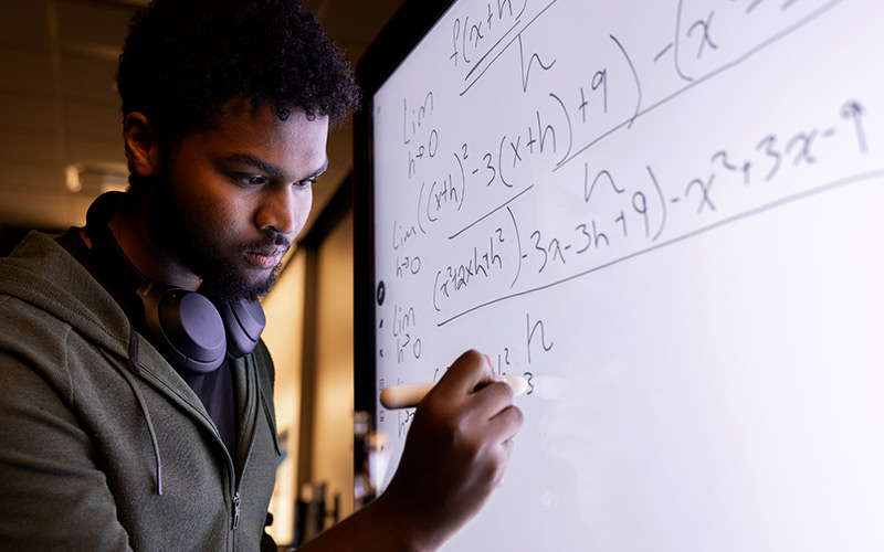 A male student holds a digital pencil to a screen as he writes out equations on a digital white board.