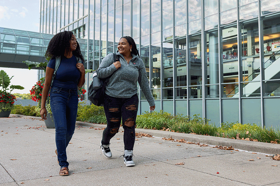 Two students walking outside of the IU Indianapolis Campus Center on a sunny day in the early fall. Both are carrying backpacks while smiling and talking to each other.