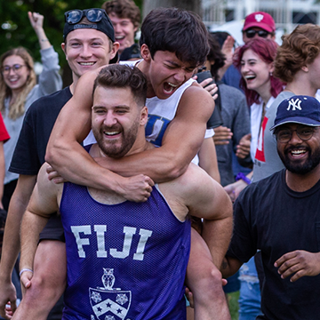 A crowd of students joyfully participates in the FIJI fraternity event. Among them, one male student is cheerfully carrying another on his back, creating a fun and spirited atmosphere, with smiles all around.