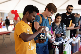 Students line up in a tent to add topics to their ice cream during the annual ice cream social. The student most prominent is wearing a yellow shirt and is adding sprinkles to his ice cream with whipped cream.