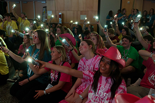 A crowd of about 20 students, mostly female, look forward at the stage during Jagathon. They are holding up cell phones their flashlights pointed toward the stage. 