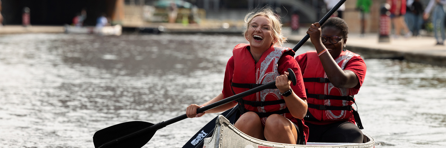 Two students are joyfully paddling their canoe during the IU Indianapolis Regatta. Both of them are wearing crimson colored shirt along with life jackets.
