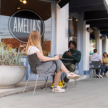 Students are gathered in front of Amelia's Bakery, enjoying their time together. The focus is on two students who are savoring their coffee while talking with each other.