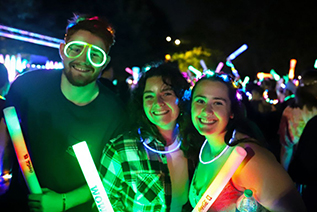 Three students pose for a picture during the Annual Weeks of Welcome event on the IU Indianapolis Campus. The male on the left is wearing neon glow-in-the dark tubes in the shape of glasses, and the two female students are wearing glow-in-the-dark necklaces. All three are holding lit up rally sticks made of foam.