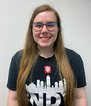 IU Indianapolis ambassador Courtney stands in front of a white wall and is wearing glasses and a black shirt that has the Indianapolis skyline and says INDY in big block letters.