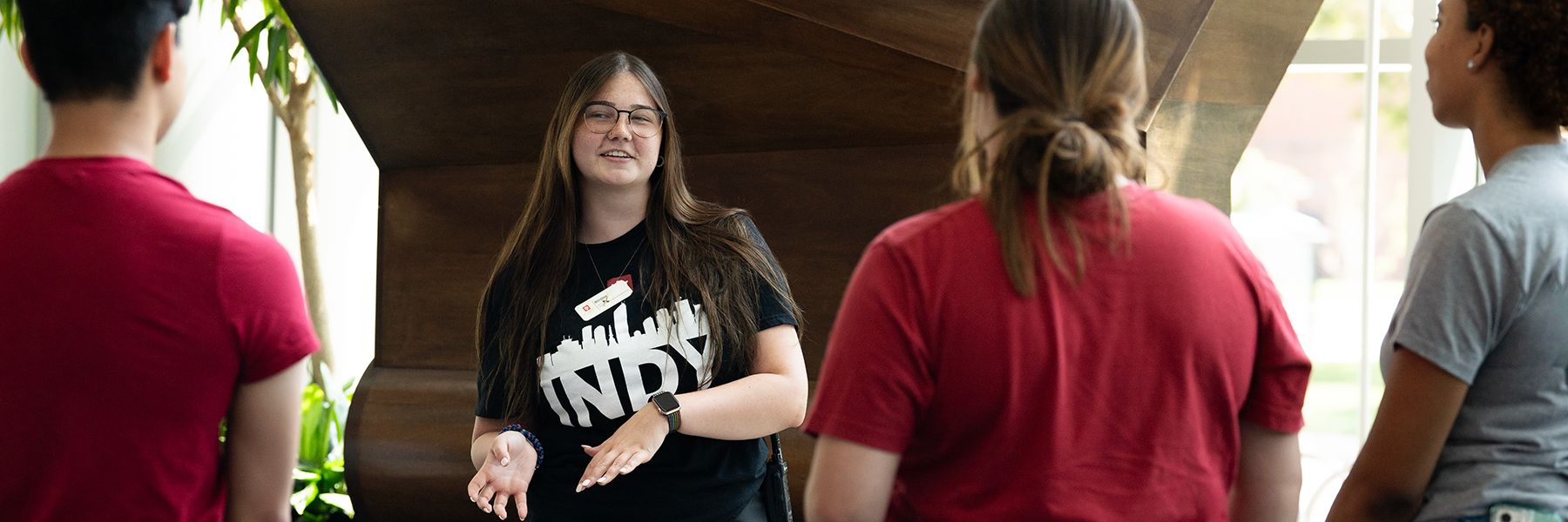 An IU Indianapolis student smiles and gestures with her hands while leading a tour of campus in front of the Punctuation Spire statue in the Campus Center to three prospective students who are facing her. She is wearing a black shirt that says INDY and has the IU trident in red.  The two students to the left are both wearing a red shirt, and the one on the right is wearing a grey shirt.