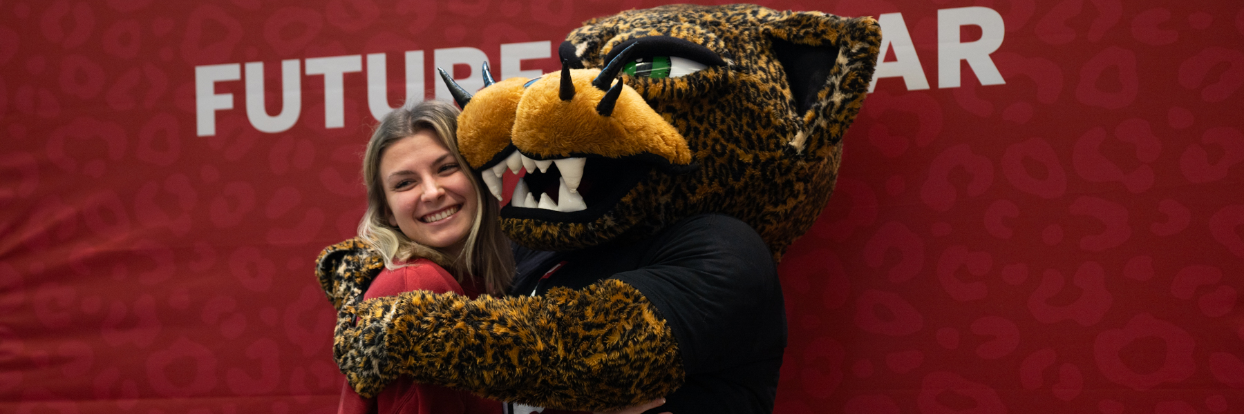 A newly admitted student poses in a hug with the IU Indianapolis jaguar mascot Jawz in front of a crimson banner that says future Jaguar and has red on red jaguar spots