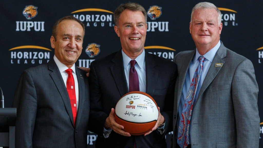 Chancellor Nasser Paydar, MayorJoe Hogsett, and Jon LeCrone stand side by side in suits in front of a banner of repeating Horizon League logos. Joe Hogsett holds a basketball with several signatures on it.