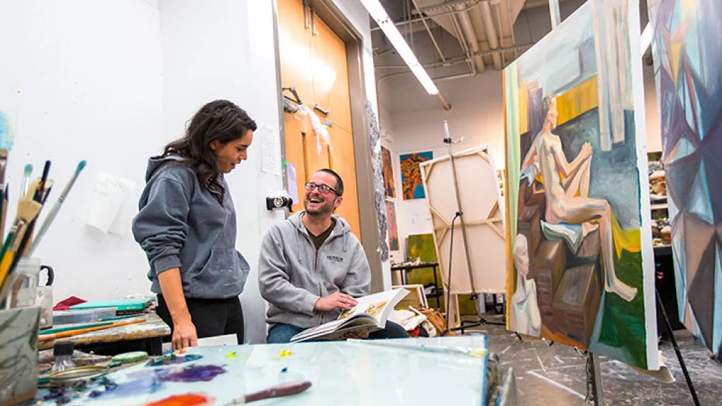 Two students stand in front of canvases, paint visible in the foreground, in Herron School of Art and Design’s painting studio.