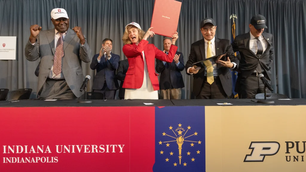 President Pamela Whitten holds up a leather folio as board of trustees members and Purdue leadership clap following the joint agreement signing ceremony that officially creates separate and independent Indianapolis campuses.