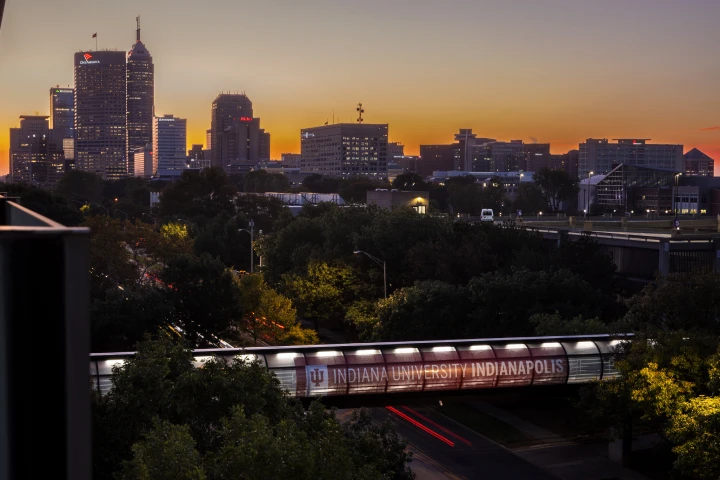 A skywalk above a street, decorated with an IU Indianapolis banner, is seen in the foreground as the sun rises in the distance behind the Indianapolis skyline.