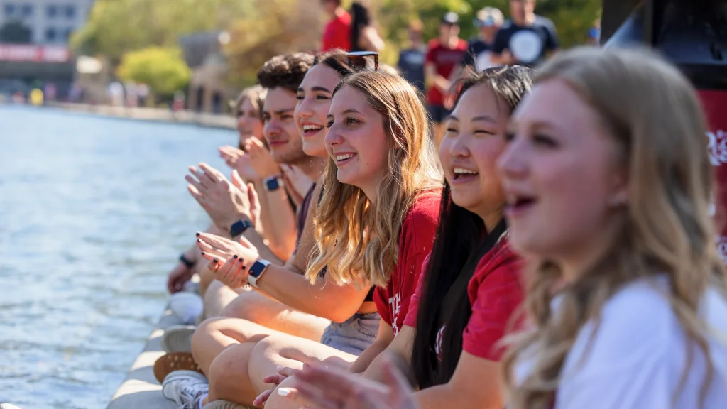 Students sit along the edge of the downtown Indianapolis canal clapping for participants in the Regatta competition.