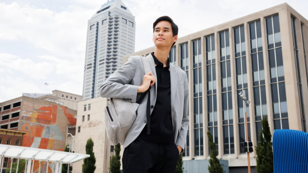 A student wearing black with a gray sport coat stands holding a backpack, downtown Indianapolis buildings and public artwork behind him.