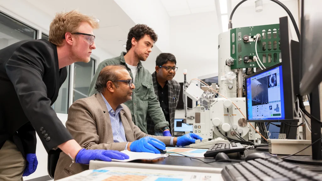 A professor is surrounded by three students looking at screens near lab equipment at the Integrated Nanosystems Development Institute.