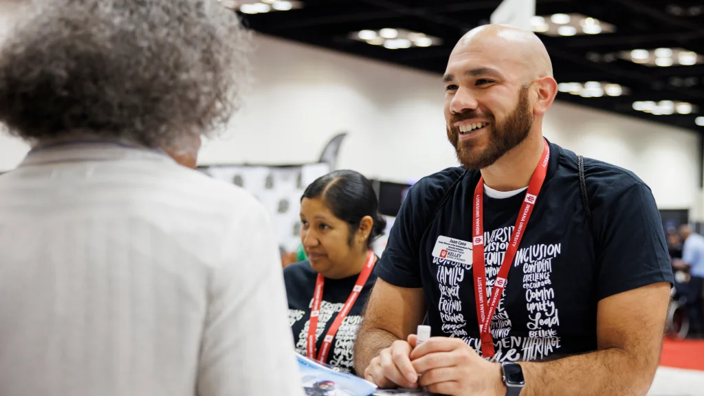 An IUI staff member wearing an IU shirt and lanyard stands at a conference table speaking with an event attendee.