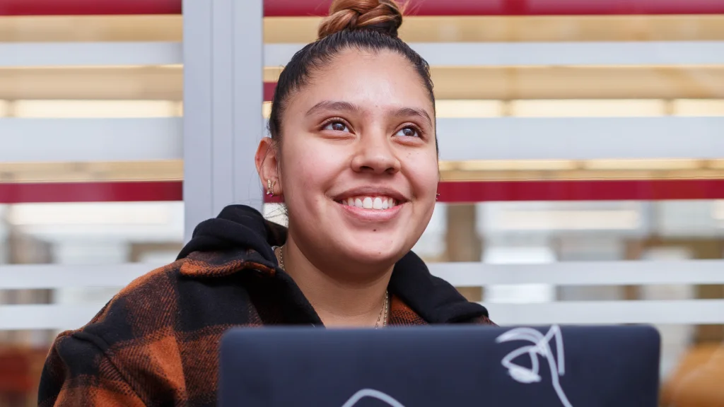 A student sits in front of a glass wall with horizontalwhite and red stripes, a laptop on her lap; she is looking up and smiling.