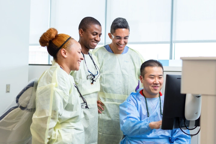 Three dental students in yellow scrubs stand around a seated instructor in blue scrubs pointing to a screen.