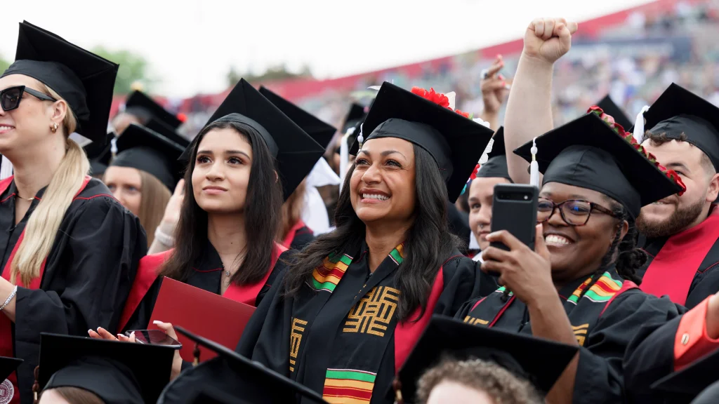 Students stand smiling at a Commencement ceremony; ones holds up a phone to take an image, another behind her holds an arm in the air in excitement.