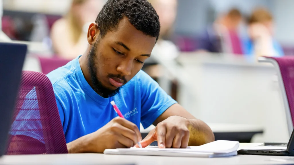 A male student writes in a notebook curing class; more student seen out of focus in rows behind him.