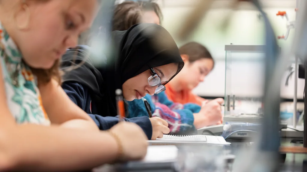 A row of four students lean over notebooks on a chemistry lab counter.