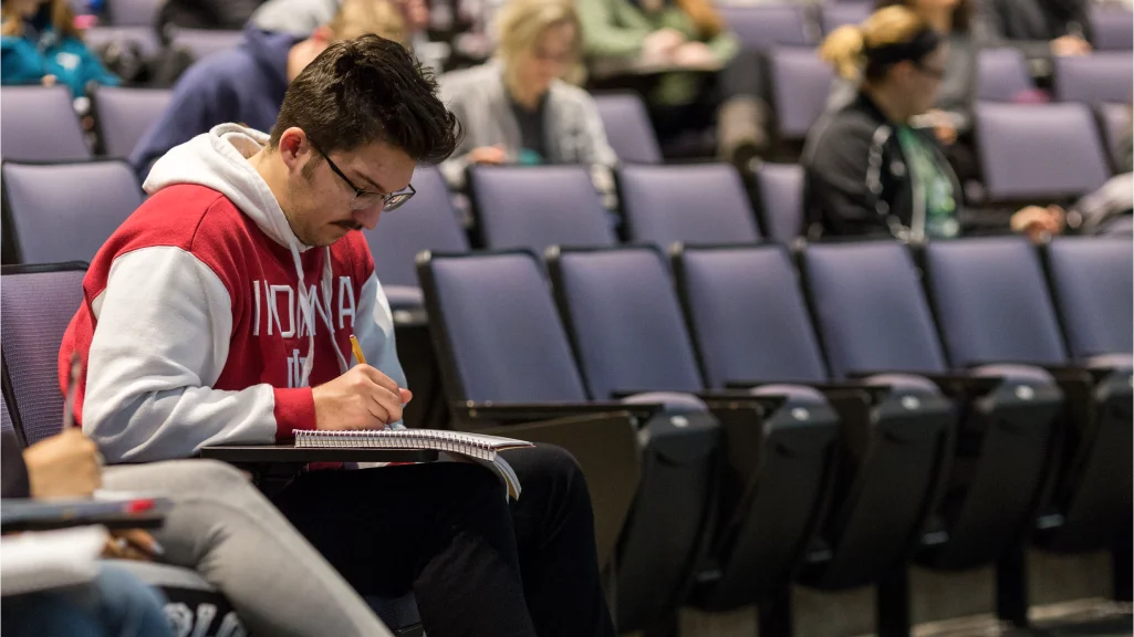A man in an Indiana sweatshirt sits in the front row of classroom seats writing in a notebook.