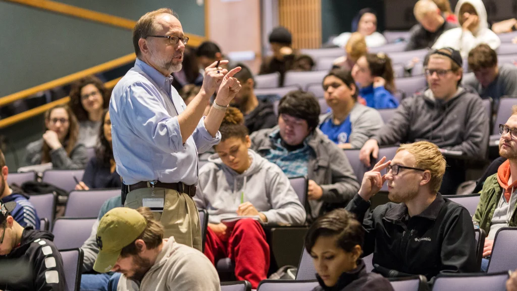 An instructor stands in the aisle of a lecture hall speaking and gesturing with his hands while students watch or take notes during the class.
