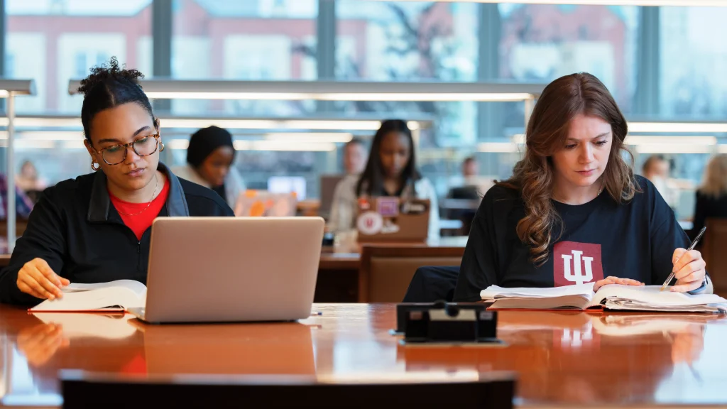 Two students sit at a long wooden table with textbooks open in front of them. Additional tables with more students extend into the background.