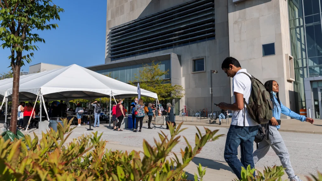 Two students pass by walking outside the campus center. A white event tent is set up in the street  to the left for an event and landscaping is seen in the foreground.