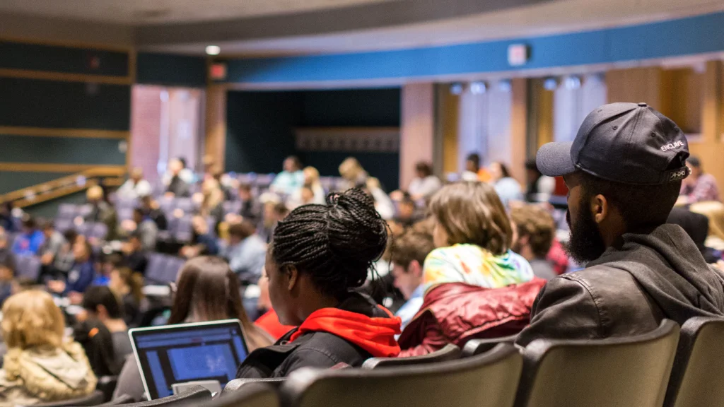 View over students’ shoulders as they sit in curved rows of chairs in a lecture hall for class.