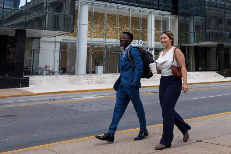 Two students in professional wear walk down a sidewalk in downtown Indianapolis, a glass-paneled building entrance seen in the background.