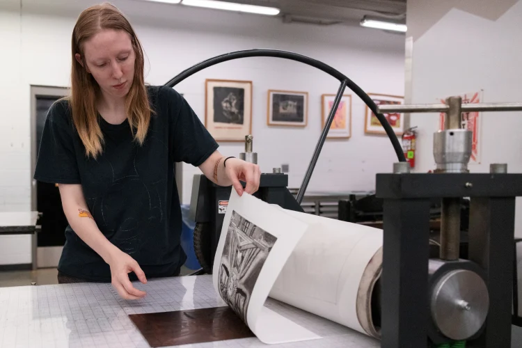 A student pulls up a pieces of artwork on paper that has been inked from an etching. The etching sits on a table with a heavy roller press with large circular handle.