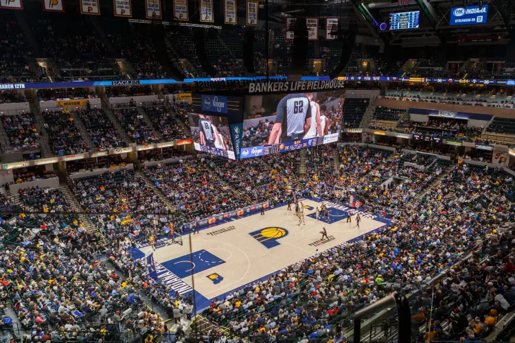 Basketball teams play on court in a packed arena. The Indiana Pacers logo is visible on center court and a large hanging screen display is centered looking down from a high level within the arena.