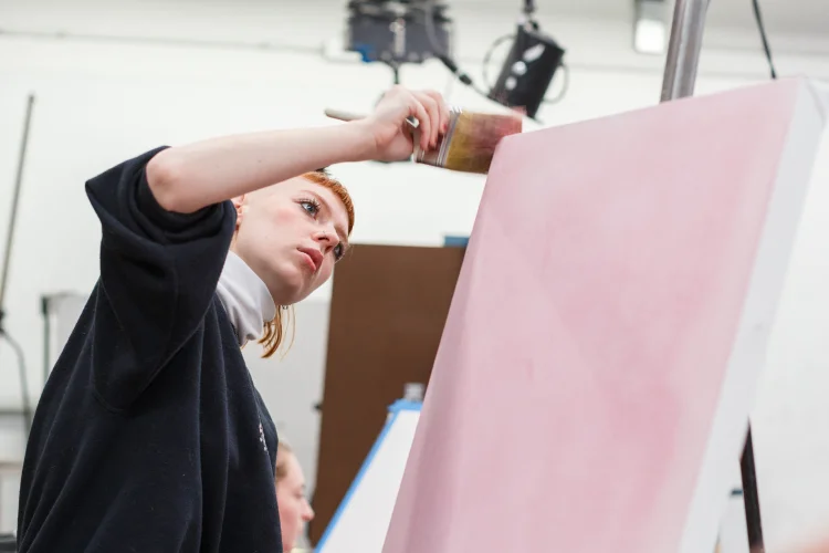 A student in a large studio paints the edge of a large canvas.