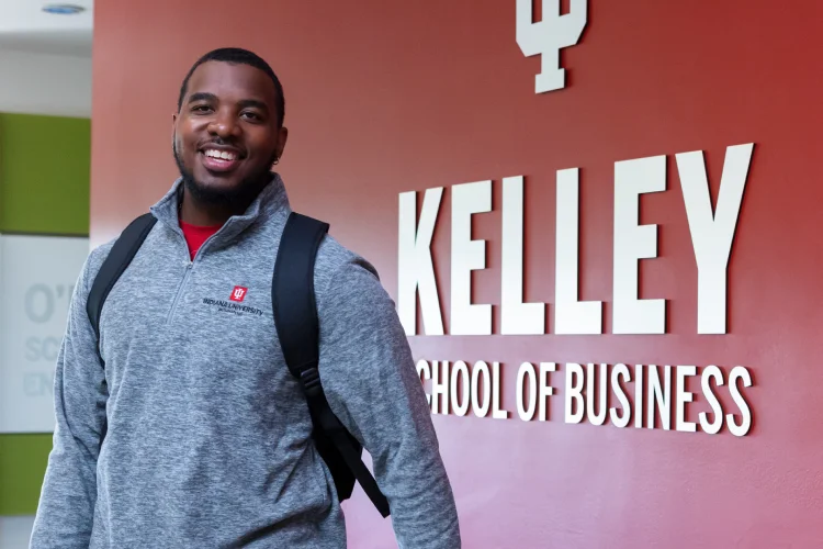 A confident, bearded young man in a gray zip-up, black backpack, and jewelry walks through the Kelley School of Business. 