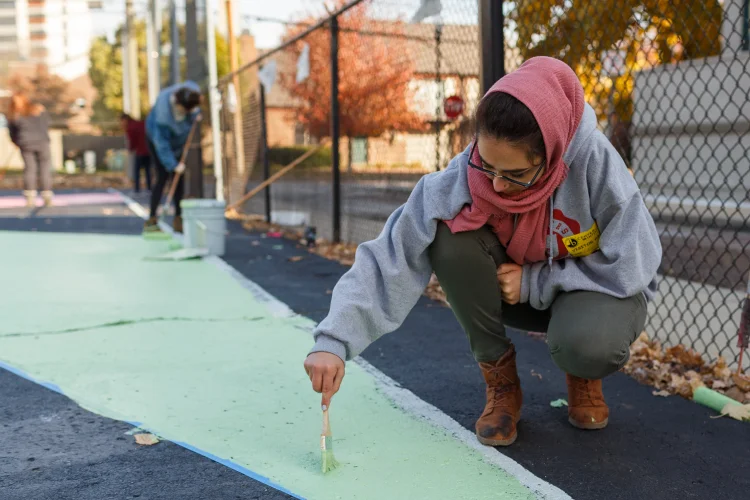 A young woman in work boots, sweatshirt, and a pink scarf paints a community mural alongside a small group.