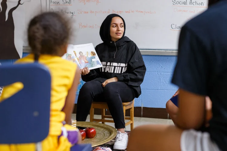 A young woman wearing modest dress reads a picture book to young children in class.