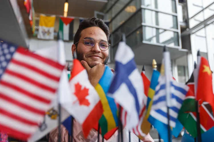 A bearded young man with glasses smiles surrounded by brightly colored table flags of the world.