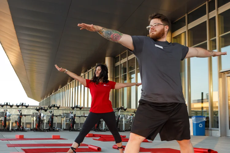 A young man in athletic wear, glasses, and tattoos practices yoga with an instructor at the Campus Center.