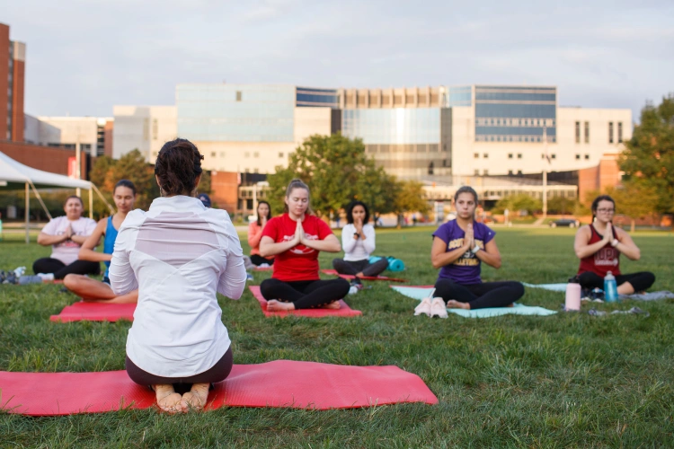 A group of about eight students spread out on a grassy lawn on campus with yoga mats. They stand with hands pressed together in front of them.