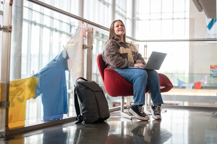 A student sits on a red upholstered chair, backpack on the ground nearby, with a laptop in her lap as she looks up and smiles.