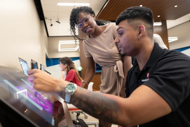 A woman leans over the shoulder of a male working on a tilted screen monitor in a classroom or computer lab area.